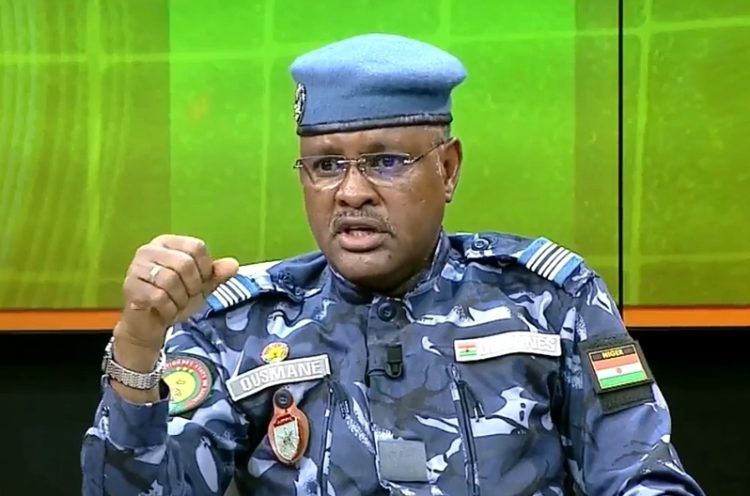 Portrait en gros plan du Colonel Mahaman Elhadj Ousmane, en uniforme militaire bleu, portant des lunettes et un béret, s'exprimant avec un geste de la main lors d'un plateau télévisé sur fond vert.