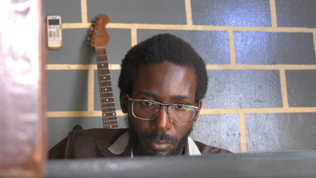 Portrait serré en contre-plongée de Mahaman Sani HOUSSEYNI ISSA (Mog), un développeur noir portant des lunettes de vue et une barbe. Il regarde vers le bas avec une expression de profonde concentration. En arrière-plan, on aperçoit un mur de briques grises aux joints clairs et le manche d'une guitare électrique en bois.
