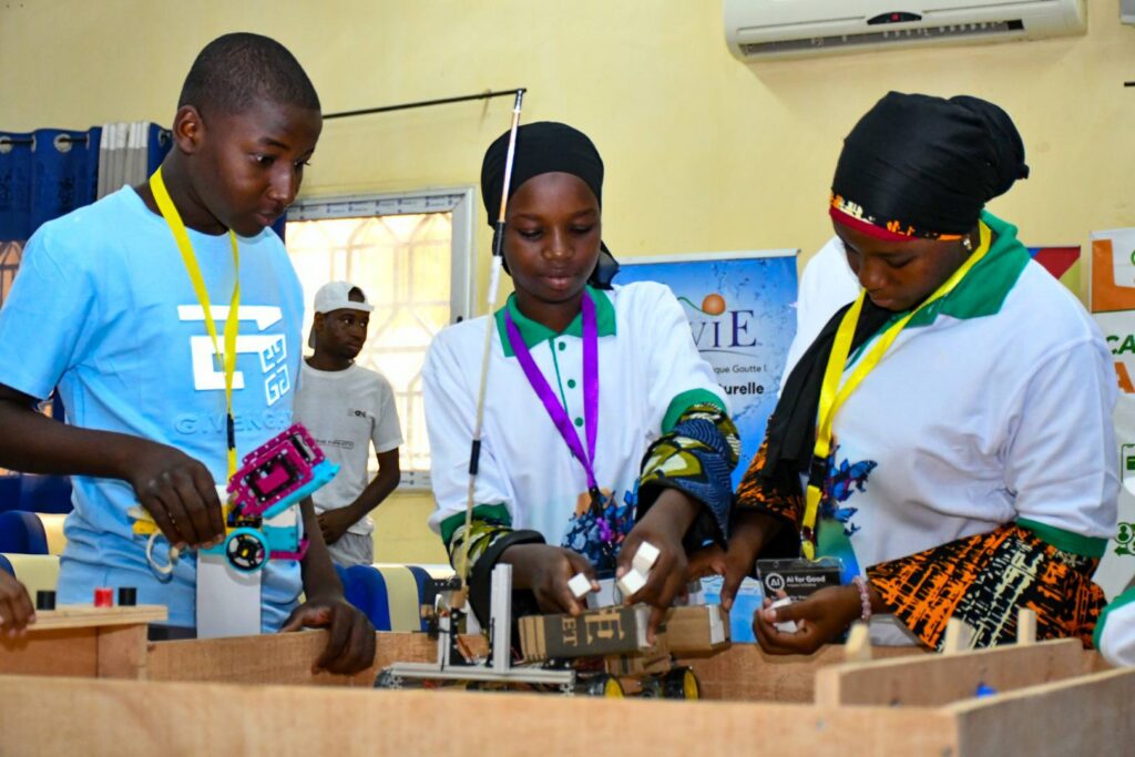 Trois jeunes participants nigériens, deux filles en blanc et un garçon en bleu, assemblent et programment un robot éducatif sur un plateau de jeu en bois lors d'une compétition de robotique.