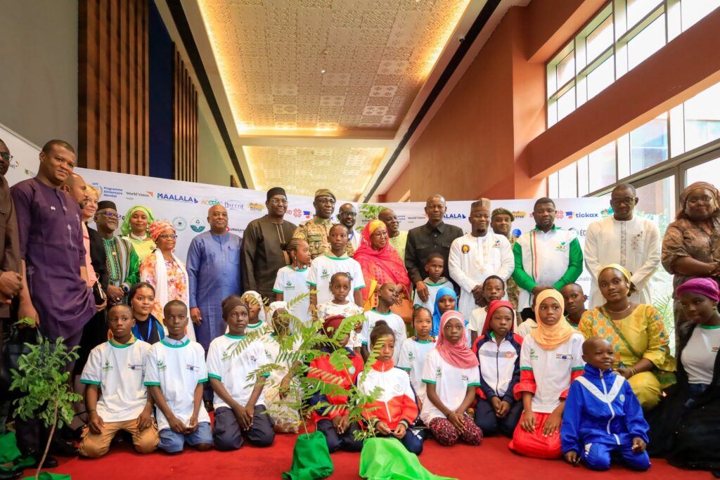 Une photo de groupe réunissant des adultes et des enfants lors du Forum national de l'Arbre à Niamey. Au premier plan, de nombreux enfants assis autour d'un jeune arbre en pot symbolisent la jeunesse et l'avenir écologique. Derrière eux, un groupe d'officiels, de dignitaires, de militaires et d'experts, hommes et femmes, se tient debout devant un mur de logos de partenaires. L'ambiance est officielle et collective, soulignant l'engagement pour l'environnement.