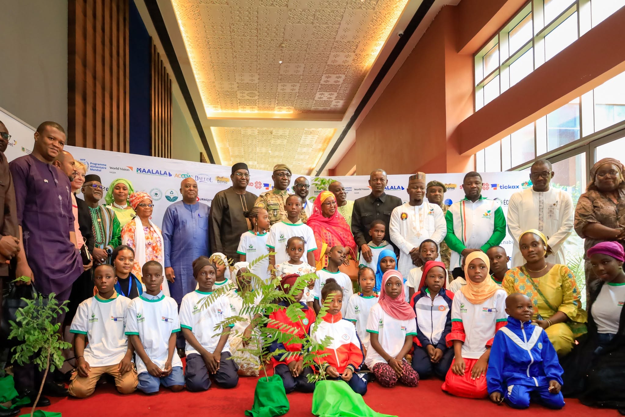 Une photo de groupe réunissant des adultes et des enfants lors du Forum national de l'Arbre à Niamey. Au premier plan, de nombreux enfants assis autour d'un jeune arbre en pot symbolisent la jeunesse et l'avenir écologique. Derrière eux, un groupe d'officiels, de dignitaires, de militaires et d'experts, hommes et femmes, se tient debout devant un mur de logos de partenaires. L'ambiance est officielle et collective, soulignant l'engagement pour l'environnement.